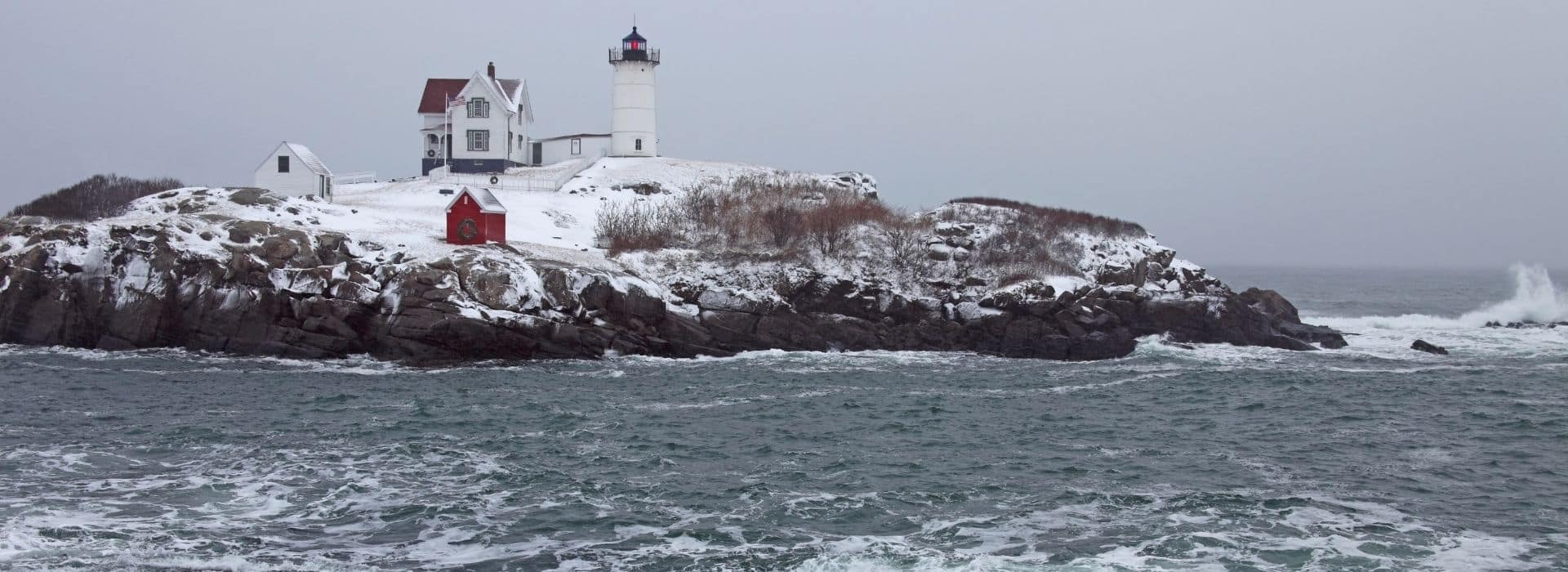 view of cape neddick on nubble on a gray wintry day with grounds covered in snow and the sea churning up