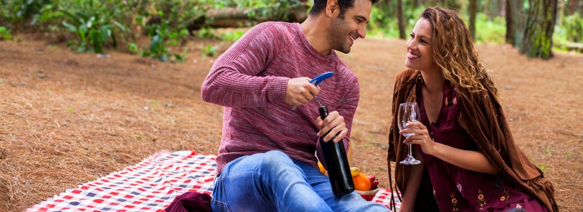 a couple enjoying a picnic in the woods. the man is opening a bottle of wine as the couple sits on a blanket.