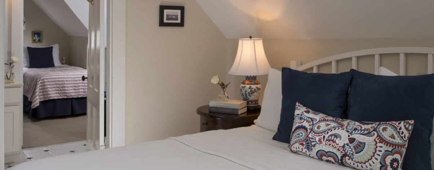 Bedroom with light cream walls, white trim, white headboard, white and navy bedding, and view of twin bed in adjoining room