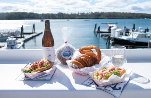 White counter with lobster rolls, fried onion rings, bottle of beer, and view of a marina