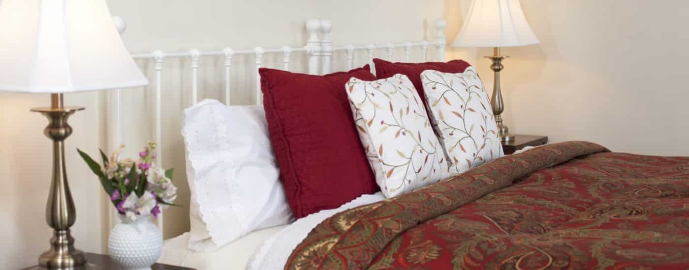 Bedroom with white walls, white painted wrought iron headboard, red and gold paisley bedding, and dark wooden furniture
