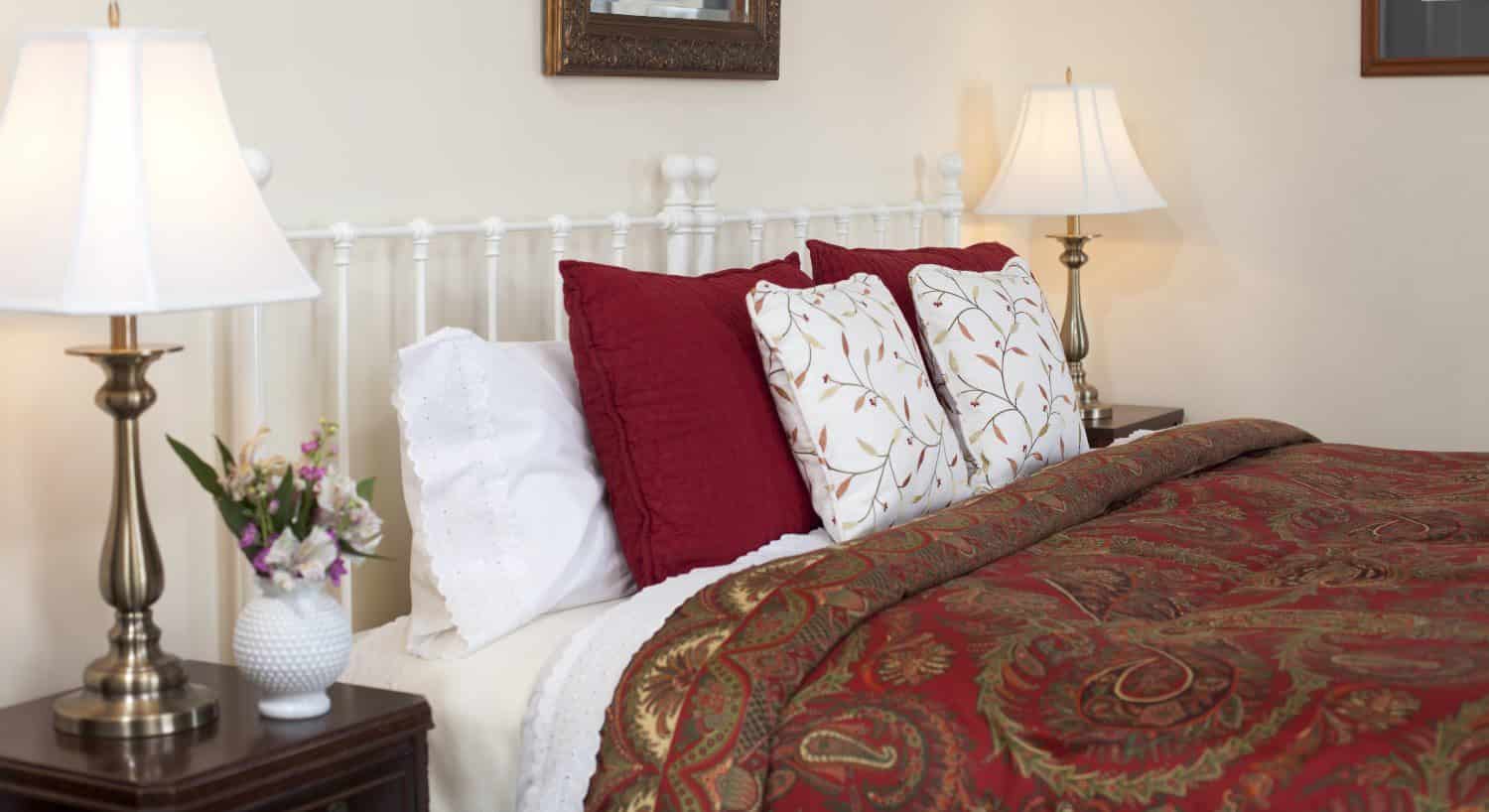 Bedroom with white walls, white painted wrought iron headboard, red and gold paisley bedding, and dark wooden furniture