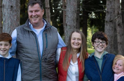 Man in gray vest, woman in red vest, two boys in blue vest, and girl in pink vest standing next to each other with trees in the background