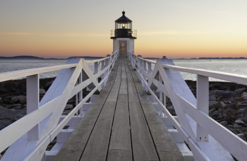 Small lighthouse at the end of a long boardwalk with white fencing on both sides and ocean in the background at dusk