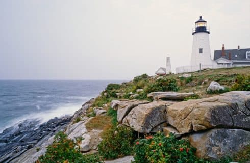 Large white lighthouse on a hill surrounded by large rocks with large waves coming in