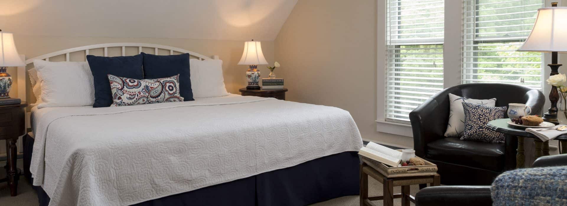 Bedroom with light cream walls, white trim, white headboard, white and navy bedding, sitting area, and large windows