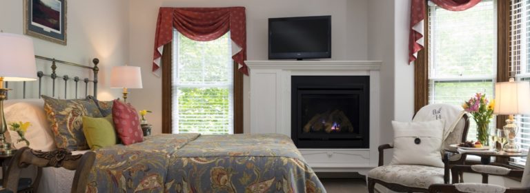 Bedroom with light-colored walls, dark wooden trim, blueish gray painted wrought iron headboard, multicolored bedding, and fireplace